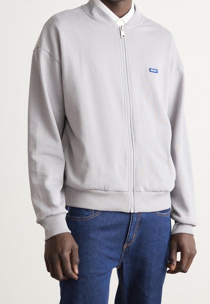Mand iført en lys grå zip-up bomberjakke med et lille blåt "HUGO" logo og blå jeans, der står op ad en ensfarvet baggrund.