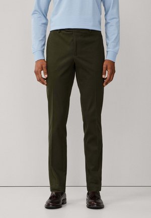 Man draagt strakke donkerolijfgroene broek, lichtblauw overhemd met lange mouwen en donkerbruine loafers met kwastjes, staand voor een effen achtergrond.