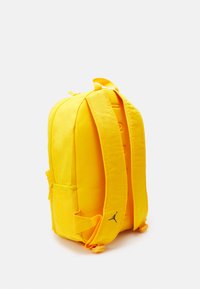 Zaino giallo in tessuto con spallacci imbottiti e scomparto principale con zip. Presenta una tasca laterale e un dettaglio del logo nero.