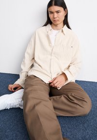 Cremfarbendes, gestreiftes Hemd mit Brusttasche, kombiniert mit lockeren braunen Hosen und weißen Sneakers, auf einem blauen Teppich sitzend.