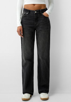 Personne portant un jean noir taille haute à jambes larges, un crop top blanc à manches longues et des baskets blanches, debout avec une main dans la poche.