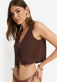 Καφέ κοντό τοπ με V-λαιμόκοψη και δέσιμο μπροστά. Το μοντέλο φορά σπείρες σκουλαρίκια και βραχιόλια. Απαλή, ελαφριά υφή.