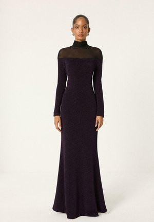 Femme portant une longue robe ajustée violet foncé avec un col montant noir transparent et des manches longues, debout devant un fond clair uni.