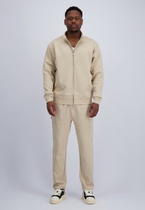 Mann steht und trägt eine beige Jacke mit Reißverschluss, passende beige Hose, weißes Unterhemd und schwarze weiße Sneaker vor hellem Hintergrund.