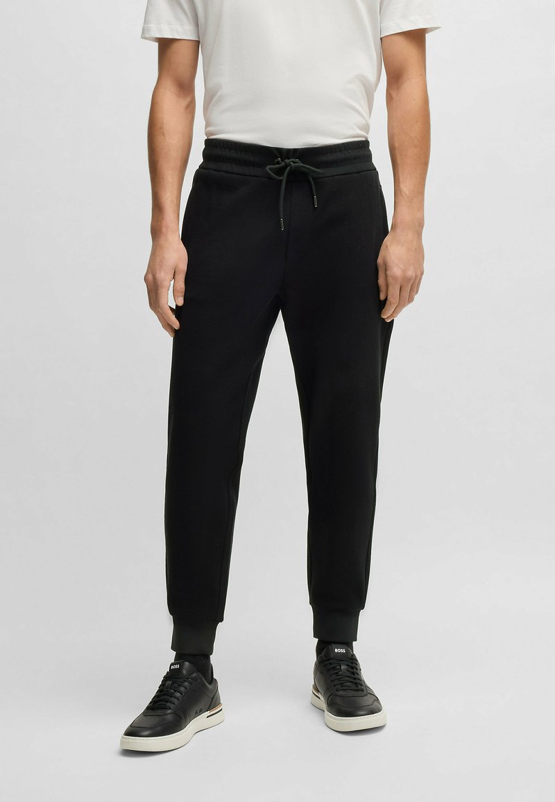 Svarta bomullsbyxor med elastisk midja och justerbar dragsko, nedtaperad bendesign och ribbade mudd, tillsammans med svarta sneakers.