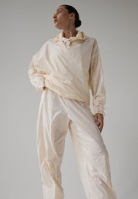 Kobieta stojąca z rękami na biodrach, ubrana w luźną, kremową kurtkę z kapturem i pasujące spodnie na jednolitym tle.