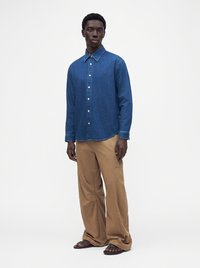 Homme portant une chemise bleue à manches longues boutonnée, un pantalon beige ample et des sandales foncées, debout devant un fond blanc uni.