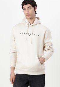 Crème kleurige hoodie gemaakt van zachte stof met een voorvak. Voorzien van een logo "TOMMYJEANS" in het zwart over de borst.