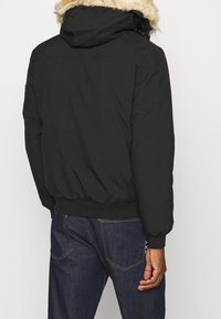 Veste matelassée noire avec une capuche doublée de fourrure, des poignets et une taille élastiques. Le dos présente une finition lisse et est associé à un jean bleu foncé.