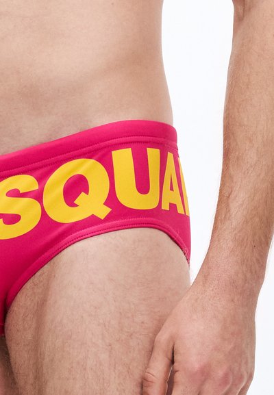 Rožinės maudymosi glaudės su plačia geltona juosmens juosta, ant kurios parašyta „SQUAD“. Audinys lygus, glaudžiai aptempia, pabrėždamas kūno formą.