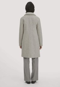 Grauer Wollmantel mit strukturiertem Kragen, langen Ärmeln und geradem Schnitt, von hinten gezeigt. Kombiniert mit grauer Hose und flachen Schuhen.