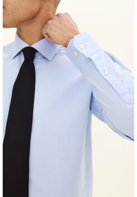 Ljusblå skjorta med klassisk krage, långa ärmar och vita knappmanchetter. Bärs med en svart slips som visar en slät yta.