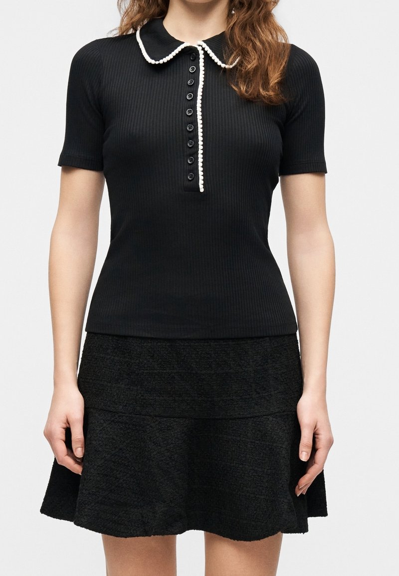 Femme portant un polo noir ajusté à manches courtes côtelé avec une bordure blanche au col et à la patte de boutonnage, associé à une jupe noire texturée.