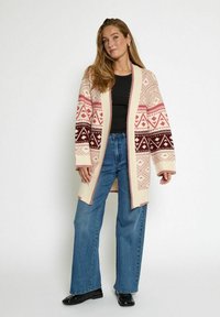 Gebreide cardigan in crème met geometrische patronen in bordeaux en roze, open voorkant, verlaagde schouders en geribbelde manchetten, gecombineerd met blauwe jeans.