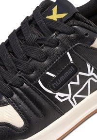 Svart lädersneaker med gräddfärgade detaljer, prydd med ett vitt geometriskt mönster, texturerad tygtunga och svarta snören. Logotypen visas.