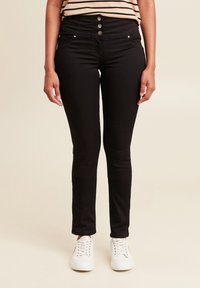 Jean noir taille haute avec une fermeture à boutons et cinq poches, associé à des baskets blanches. Le tissu est lisse avec un léger stretch.