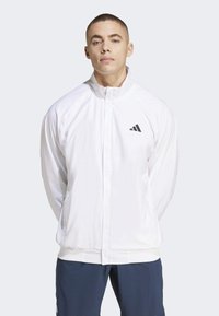 Witte sportjack met hoge kraag, ritsluiting en zwart logo op de borst. Heeft een gladde textuur en elastische manchetten.