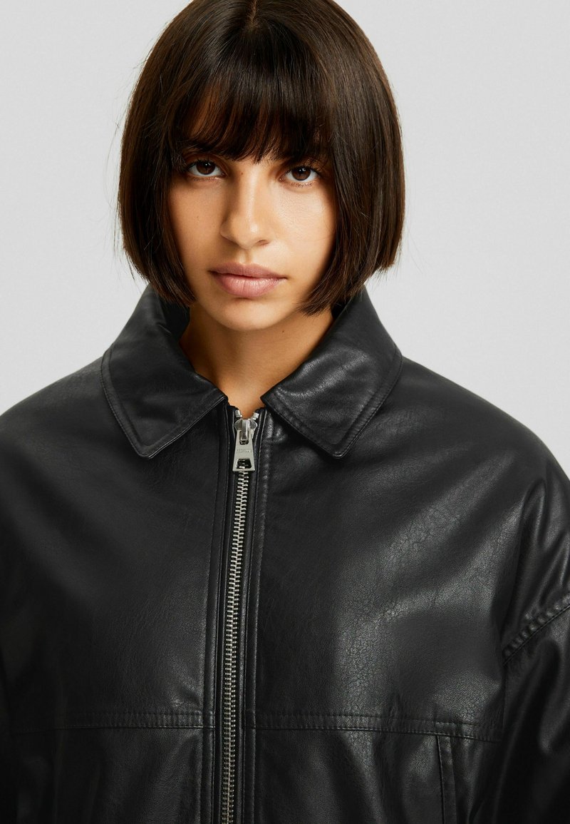 Chaqueta de cuero negra con cuello puntiagudo, cierre frontal mediante cremallera, textura suave y un leve brillo. Corte entallado con costuras en los hombros y bolsillos.