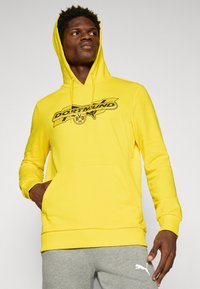 Gul hættetrøje med en front kangaroo-lomme og ribbede manchetter, med et sort grafisk print af "DORTMUND" og logo på brystet.