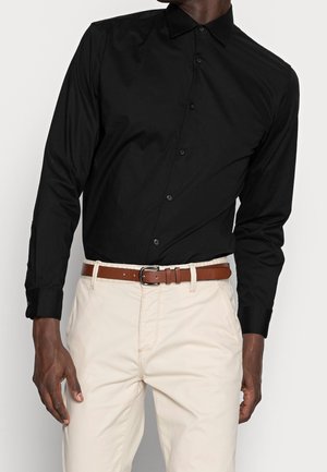 Homme portant une chemise noire à manches longues boutonnée rentrée dans un pantalon beige avec une ceinture en cuir marron, debout devant un fond uni.