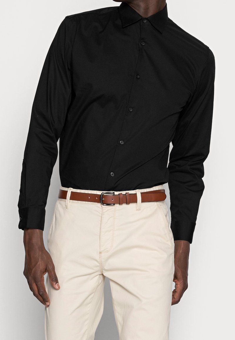 Homme portant une chemise noire à manches longues boutonnée rentrée dans un pantalon beige avec une ceinture en cuir marron, debout devant un fond uni.