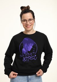 Giovane donna con occhiali e capelli raccolti a chignon indossa un felpa nera con una grafica viola del profilo di una ragazza e la scritta "Sub-Urban Gothic".