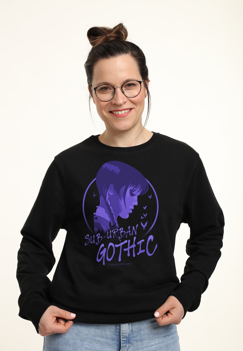 Giovane donna con occhiali e capelli raccolti a chignon indossa un felpa nera con una grafica viola del profilo di una ragazza e la scritta "Sub-Urban Gothic".