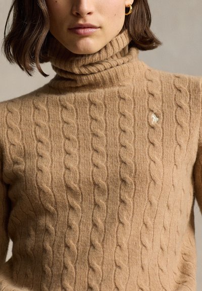 Femme portant un pull à col roulé en maille torsadée couleur chameau avec un petit logo blanc brodé sur le côté gauche de la poitrine et des créoles dorées.