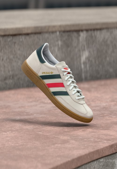 Bílá teniska Adidas se zelenými a červenými pruhy, gumovou podrážkou a nápisem „SPEZIAL“ vznášející se nad kamennou dlažbou.