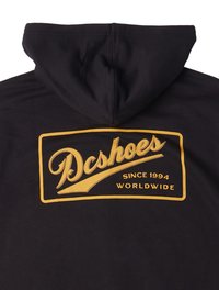 Sudadera negra con capucha que presenta un gran logo amarillo de "Dcshoes" con el texto "Desde 1994" y "Mundial" en una fuente retro. Tela suave y casual.