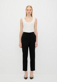 Witte geribbelde tanktop gecombineerd met op maat gemaakte zwarte broek. Model draagt zwarte puntige schoenen met gouden accenten. Eenvoudige achtergrond.