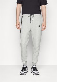 Pantaloni della tuta grigi in tessuto misto cotone, con vita elasticizzata e cordino, tasche laterali con zip e dettagli neri. Logo Nike presente.