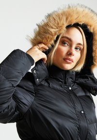 Svart puffjakke med høy krage og hette foret med kunstpels. Har trykknapper og vattert tekstur. Glatt finish.