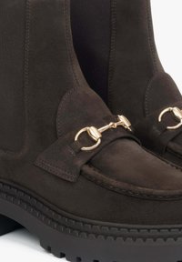 Ciemnobrązowe zamszowe botki za kostkę z elastycznymi panelami bocznymi, ozdobnymi złotymi elementami oraz solidnymi gumowymi podeszwami. Całość wykończona teksturowaną powierzchnią.