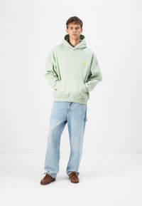 Lys grønn hettegenser med frontlomme og gule grafiske detaljer, kombinert med løse lyseblå jeans og brune båtsko.