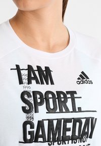 Bílé bavlněné tričko s tučným černým textem, včetně "I AM SPORT" a "GAMEDAY", s logem Adidas v pravém horním rohu.