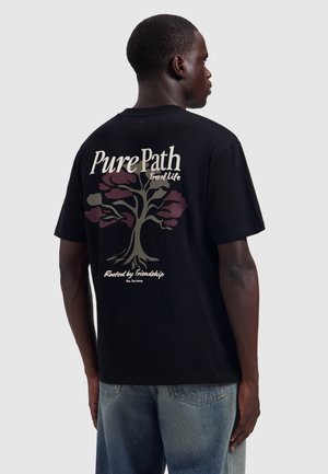 Man die van zich af kijkt, draagt een zwart T-shirt met een boomgrafiek en de tekst "Pure Path, Tree of Life, Rooted by friendship" en een blauwe spijkerbroek.