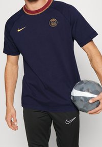 Camiseta de Nike azul marino con cuello burdeos, con un logo del Paris Saint-Germain en dorado. Pantalones negros con un swoosh blanco. Sosteniendo una pelota gris.