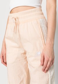 Světle broskvové lehké kalhoty s elastickým pasem, šňůrkou a vyšitým logem "New Balance" na levé straně.