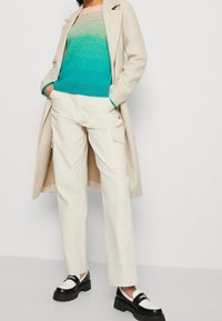 Beige vlněný kabát na vrchu teal a růžový pruhovaný pletený svetr. Směrem dolů krémové cargo kalhoty a černé platformové mokasíny s bílými akcenty dotváří vzhled.