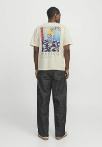Beige T-Shirt mit kurzen Ärmeln und einem bunten Matisse-Kunstwerk-Druck auf dem Rücken. Kombiniert mit dunkelblauen, weit geschnittenen Jeans und hellen Schuhen.