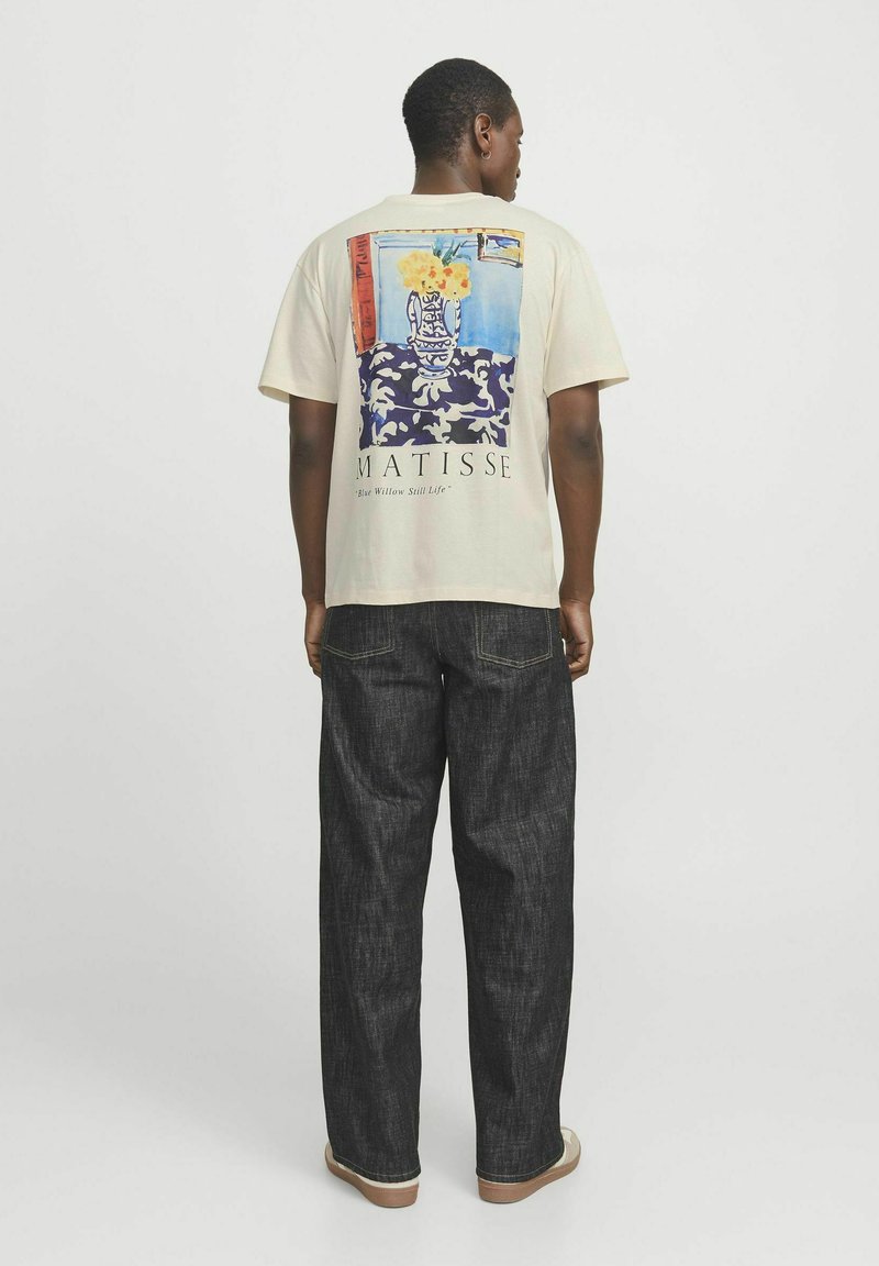 Beige T-Shirt mit kurzen Ärmeln und einem bunten Matisse-Kunstwerk-Druck auf dem Rücken. Kombiniert mit dunkelblauen, weit geschnittenen Jeans und hellen Schuhen.