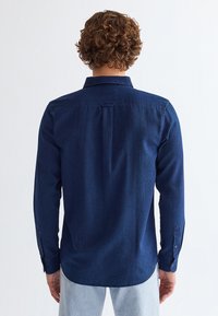 Uomo con capelli ricci che indossa una camicia blu scuro a maniche lunghe con bottoni e jeans azzurri, visto da dietro su sfondo bianco.