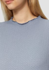 Helles blaues Oberteil aus einem strukturierten, gewebten Stoff mit dezentem hexagonalem Muster und einem rohen, abgerundeten Ausschnitt.