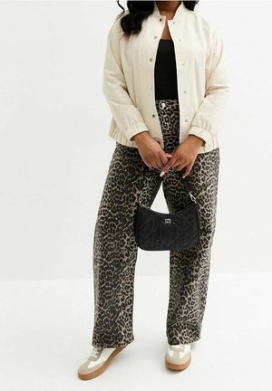 Mujer con chaqueta beige, camiseta negra, pantalones amplios con estampado de leopardo y zapatillas blancas, sosteniendo un pequeño bolso negro acolchado al frente.