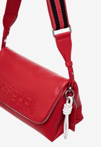 Borsa a mano in pelle rossa con finitura testurizzata, chiusura con zip e tracolla staccabile con design a strisce nere e rosse. Dettaglio hardware bianco.