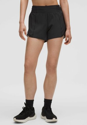 Schwarze sportliche Shorts mit hohem Bund, glatter Textur, seitlichen Schlitzen, kombiniert mit schwarzen Turnschuhen und schwarzen Knöchelsocken.