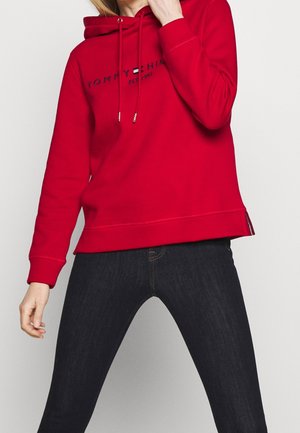 Personne portant un sweat à capuche rouge vif Tommy Hilfiger avec logo et un jean skinny foncé, debout jambes écartées et main près du visage.