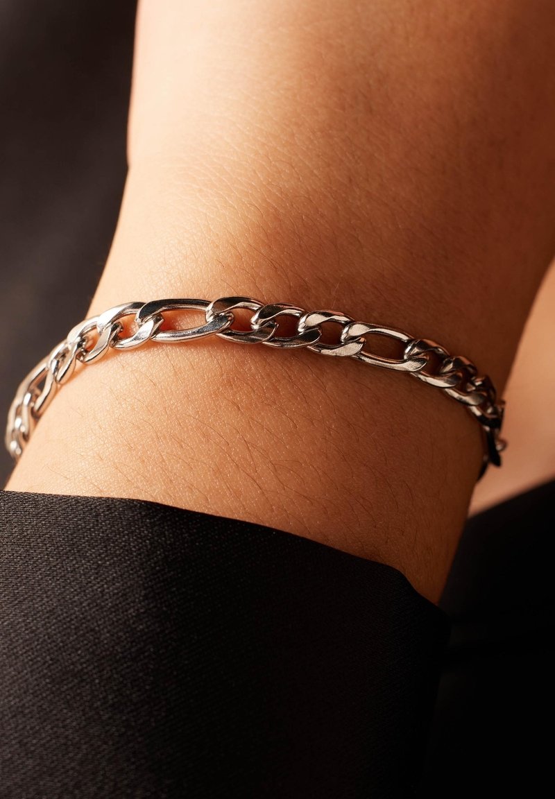 Zilveren schakelarmband om een pols, met in elkaar grijpende schakels en een gepolijste afwerking, tegen een donkere stoffen achtergrond.