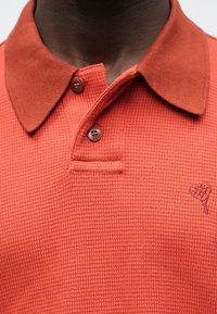 Červené polo tričko s texturou, hnědým límcem, dvěma hnědými knoflíky a malým vyšívaným srdíčkem na prsou.